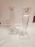 Front view of pair of tall clear crystal candlesticks with square bases and vertical ridged column design, with visible red candle wax residue in cups.
