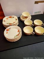 Photo showing eight 6-inch square plates with floral design and gold trim, stacked in two rows, with five teacups and four saucers in the background.