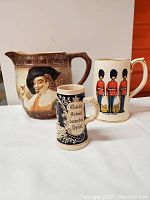 Photo showing the Falcon Ware Cavalier pitcher and two miniature beer steins side by side on a white surface with an orange background.