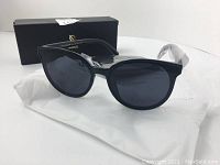 Black round sunglasses with grey lenses displayed with black branded box, white cloth pouch, and plastic wrapped small black accessory case.