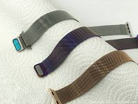 Three stainless steel mesh watch bands in silver, gold, and iridescent purple laid out on a paper towel. Showcasing texture and detailing of mesh metal and magnetic clasp ends connected to Apple Watch adapter pieces.