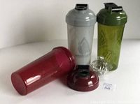 Set of three Hydra Cup shaker bottles in red, gray, and green with lids and whisk balls displayed on a round white table.