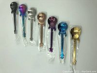 7 guitar-shaped coffee spoons in different metallic colors all sealed in individual plastic bags, laid out side by side.