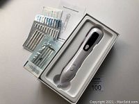 White handheld spot removal pen resting in molded white plastic tray alongside sets of replaceable needles in sealed foil pouches and an instruction manual.