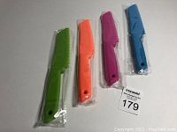 Four plastic kitchen knives in green, orange, purple, and blue, individually wrapped in plastic, placed on a white surface with a MaxSold label