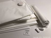 Eight white vacuum storage bags stacked and folded, showing the folded plastic with valve caps visible, alongside a rolled bag still in plastic wrap.
