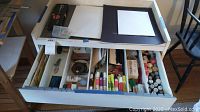 Top-down view of open large white drawer holding various art supplies separated in compartments, showing inks, chalk pastels, markers, blades, and tools