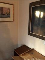 Photo showing two framed artworks hung on a wall above cardboard boxes, with reflections on glass surfaces and visible wall texture.