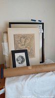 Framed artworks stacked together, showing various framed pieces including a drawing and a black and white print.