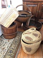 Photo showing six baskets grouped together including a wooden barrel-style basket, two large dark woven baskets with handles, a lighter square woven basket with a blue tag, and two smaller tan oval woven baskets with lids and handles featuring dark diamond patterns.
