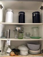 Three shelves of kitchenware including white and blue metal canisters, multiple glass bowls and pitchers, ceramic cookware, and kitchen utensils