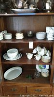 Overall view of china and serving ware arranged on wooden shelves including tureen, cups, saucers, plates, charger, and glass pieces.