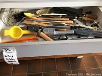 Drawer filled with various kitchen utensils and cutlery items including wooden spoons, knives, spatulas, strainer, lemon squeezer, and garlic press.