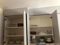 Six shelves of various plates, bowls, trivets, and small serving dishes stored inside kitchen cabinets.