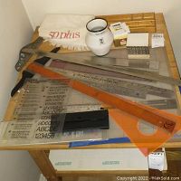 Photo shows a collection of drafting tools laid out on a wooden table including rulers, T-squares, typography guide, and a cutting mat.
