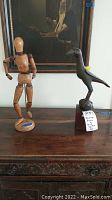 Two items on wooden table in front of framed artwork: wooden drawing figure mannequin on left and wooden bird sculpture on right with tag