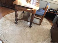 Photo shows round off-white/cream shag Karastan rug with thick pile and tassel edges under wooden table and chair, visible wear and dirt.
