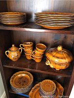 Photo of stacked plates on shelves including large and small plates, coffee cups stacked, sugar bowl with lid and a covered serving dish, all in honey glaze with floral pattern.