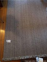 Close-up photo of the woven beige rug showing the weave pattern and fringed tassels along one edge. Minor dirt spots visible on the surface.