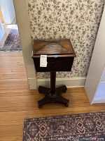 Antique wooden sewing storage box mounted on pedestal stand against floral wallpaper background placed on wooden floor with carpet partly visible. Box has a hinged lid with decorative geometric pattern and darker wood tones.