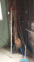 Garage corner showing long-handled garden tools including rakes, shovels, and a blue plastic broom leaning against the wall