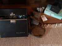 Vintage Polaroid camera carrying case with a latch lock, labeled "Basil G. Boundas" and other cameras and boxes in the background.