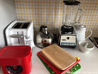 Photo of white Oster toaster, chrome tea kettle, Osterizer blender, red kitchen scale, wooden cutting boards, and white ceramic mortar and pestle on kitchen counter