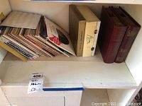 Photo shows a shelf with a loose stack of 45 vinyl records, a Bing Crosby boxed set case, and two large red binders for records.