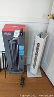 Vornado tower circulator in its original box next to the white Hitachi tower fan standing on a wooden floor.
