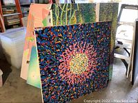 Group of paintings leaning against shelving, front view shows radial abstract piece along with additional canvases behind