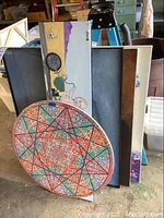 Stacked framed artworks showing various styles including a colorful circular dot-pattern geometric painting in front with partially visible figurative and abstract paintings behind.