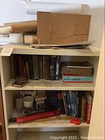 Bookshelves containing a collection of books in various sizes and conditions, along with rolled posters on top and assorted papers on lower shelves.