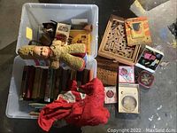View of plastic bin with vintage books, fabric soldier doll, and red jacket laid on floor next to wooden puzzles and boxed games