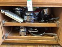 Cookware set including stainless steel pots, steamer insert, frying pan, and white baking dish inside cabinet shelves