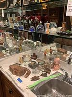 Shelf filled with glass and ceramic decorative objects including red vases, glass spheres, and assorted small items on a white cloth surface with mirror background.