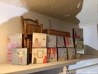 Photo showing 12 small 4x4 inch canvases with cartoon style art and humorous captions arranged on a shelf with wooden furniture background