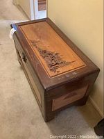 View of top and side of wooden cedar trunk showing dark wood frame and lighter inset panel with carved scenic panel featuring pagoda and people.