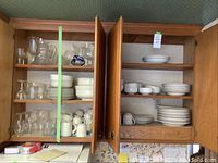 Open kitchen cabinet showing top shelves of clear glassware (pitcher, stemware) and stacked white porcelain plates, bowls and cups.