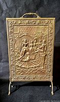 Front view of antique Victorian brass fireplace screen featuring embossed royal court scene with figures dressed in period attire, standing on curved legs