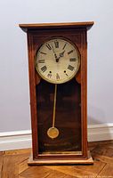 Front view of antique pendulum wall clock showing clock face with Roman numerals and brass pendulum behind glass door.