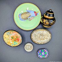 Overview of six decorative items on dark surface, showing green dish, black and gold urn, three small boxes, and enamel elephant figurine.