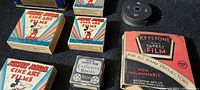 Photo showing five Mickey Mouse Cine Art Films boxes, Keystone Safety Film box, black plastic reel spool and Ramco film storage tin, on a black cloth background.