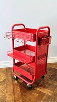Red rolling cart shown angled front side, displaying top shelf, hooks, and side basket