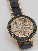Front view showing the round gold-tone watch case with black faceted bezel and crystal-encrusted swirling pattern on the face.
