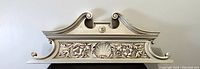 Front view of the carved decorative pediment showing symmetrical scrollwork, central shell design and floral elements with a light cream finish.