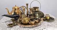 Full view of brass collection including kettle, pitchers, genie lamp, tray, and assorted vessels arranged on lace tablecloth.