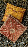 Yellow and red embellished square cushions on a patterned carpet. Yellow cushion with leaf embroidery and red cushion with intricate vine embroidery showing size and detail.