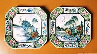 Top view of two hand painted square porcelain plates side by side showing detailed mountain and nature scenes with colorful floral borders.