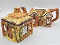 Ceramic teapot and sugar bowl resembling cottages with thatched roofs and detailed windows and doors.