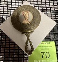 Antique hand mirror with ornate metal frame and painted portrait, displayed on a white gift box.